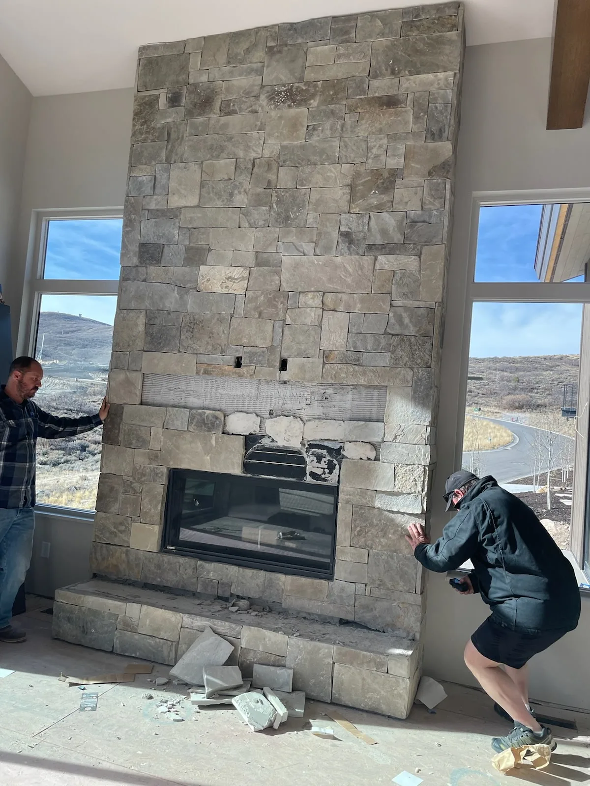 Before: a floor-to-ceiling stone veneer fireplace surround with a glass insert in a vaulted-ceiling great room, before demolition