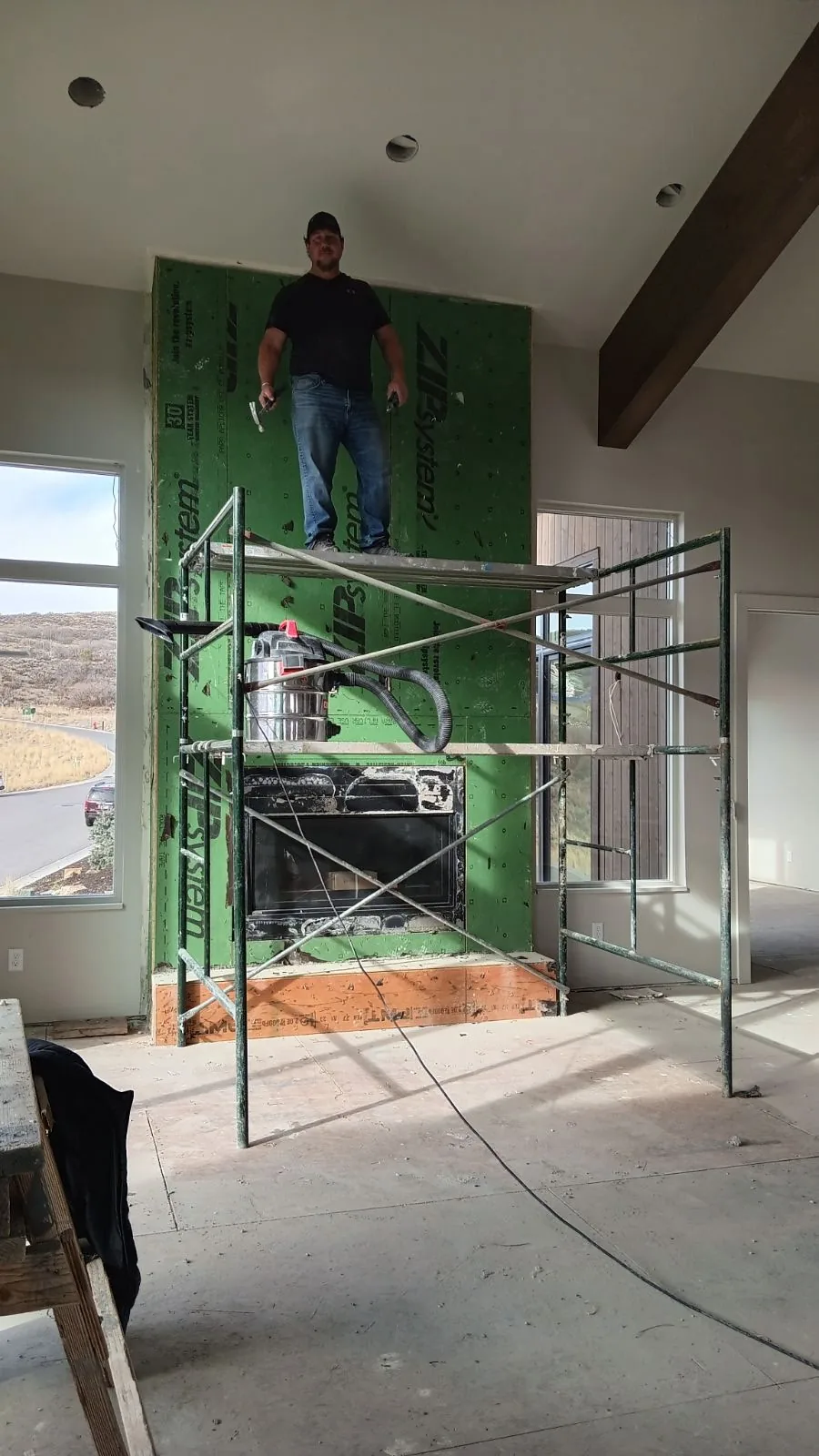 After: the same fireplace with the stone removed down to green ZIP sheathing, with a crew member standing on scaffolding